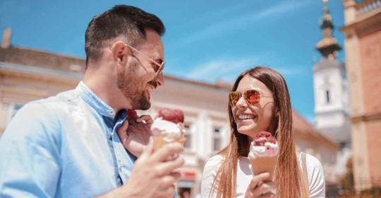 A man and woman standing outside eating ice cream cones together. They have big smiles on their faces